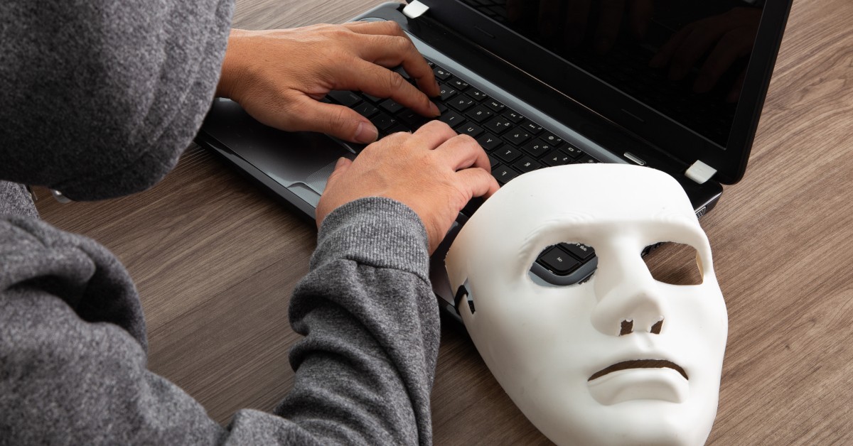 A hacker types on a computer while sitting with a mask, symbolizing the act of impersonating someone to commit an act of social engineering.